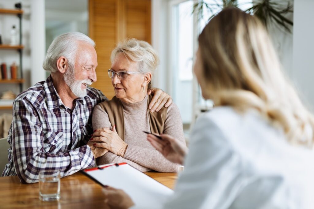 Two older adults sitting together at a table, speaking with a professional holding a clipboard in a cozy, well-lit room in the U.S.